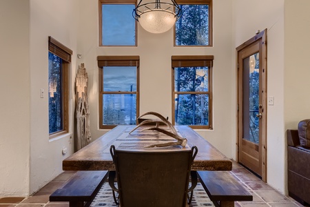 A rustic dining room with tall windows, a chandelier, a wooden table set with antlers, and a chair. The room has a natural, outdoor view through the windows and a wooden door to the right.