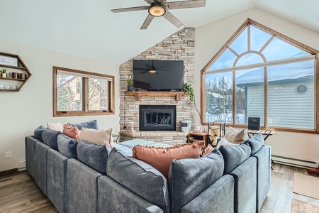 A spacious living room features a large gray sectional sofa, a stone fireplace with a mounted TV above, and large windows providing natural light and a view of the snowy outdoor scene.