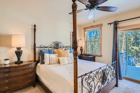 A bedroom with a four-poster bed, two bedside tables with lamps, a ceiling fan, and a window and glass door overlooking a forested area.