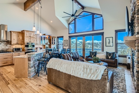 A spacious, modern living room with wooden floors, a high ceiling, large windows with mountain views, a stone fireplace, and a mix of leather and upholstered furniture.