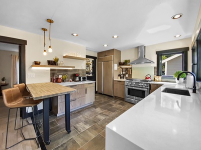 Modern kitchen with stainless steel appliances, white countertops, wooden cabinetry, and a tiled floor. There's a breakfast bar with two stools, hanging pendant lights, and a window with outdoor view.