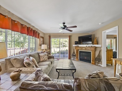 A cozy living room with beige walls, a ceiling fan, a fireplace with a TV above, and large windows with red curtains. There are two beige sofas, a coffee table, and a glimpse of a patio outside.