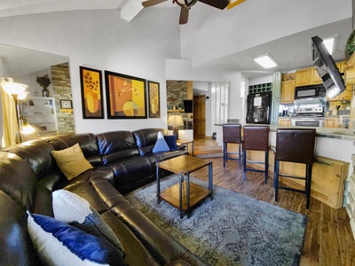 A living room with a curved black leather sectional sofa, a wooden coffee table, three bar stools at a kitchen counter, wall art, and a ceiling fan. The kitchen features stainless steel appliances.