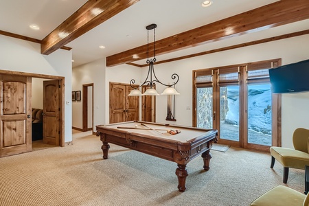 Room with a wooden pool table under a chandelier, carpeted floor, wooden beams, and doors leading outside. Snow is visible through the glass doors.