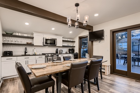 Modern dining area with a wooden table set for six, adjacent to a kitchen with white cabinets and a mounted TV. Glass doors on the right lead to an outdoor patio with more seating.