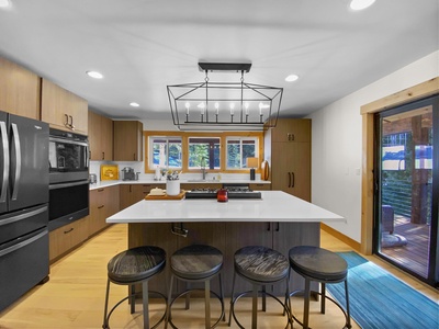 A modern kitchen with wooden cabinets, a large island with a white countertop and four stools, a dark metal chandelier, stainless steel appliances, and a sliding glass door leading to a wooden deck.