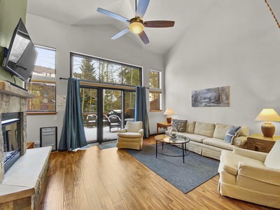 A spacious living room with a high ceiling, light tan sectional sofa, coffee table, two chairs, a wall-mounted TV, a fireplace, and large windows with blue curtains overlooking a balcony.