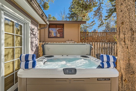 Outdoor hot tub with bubbling water and three rolled blue and white striped towels on the edge, located next to a house and a wooden fence with trees in the background.