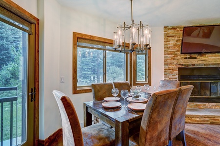 A dining room features a wooden table set with plates and glasses, surrounded by six upholstered chairs. Large windows and a glass door provide a view of greenery. A chandelier hangs above, and a TV is mounted above a stone fireplace.