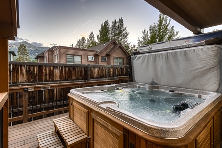 A hot tub on a wooden deck, enclosed by a wooden fence, with trees and a house in the background. The hot tub cover is partially open.