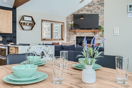 A dining table set with mint green dishes and clear glasses, a small vase with lavender flowers in the center, in a modern living-dining area with a stone fireplace and TV in the background.