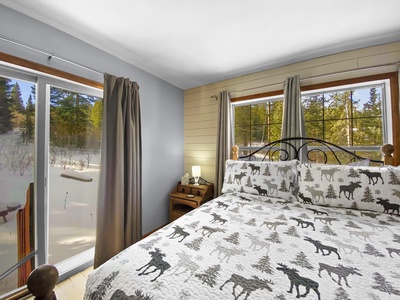 A bedroom with a large window and sliding glass door, featuring a bed with animal-themed bedding, a nightstand with a lamp, and curtains. Snow-covered trees are visible outside.