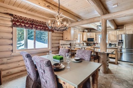 A log cabin kitchen and dining area with wooden beams, a chandelier, a set dining table, and a snow-covered window view.