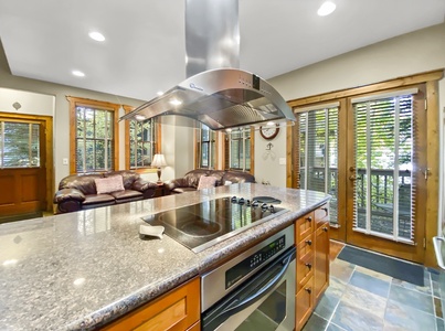 A modern kitchen with a granite countertop island, electric stovetop, and a stainless steel range hood. The kitchen opens to a living room with leather sofas and large windows with wooden blinds.