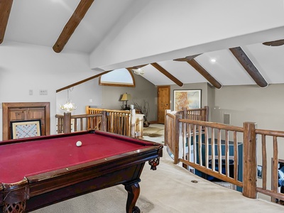 A spacious loft with exposed wooden beams features a red pool table and wooden railings, overlooking a well-lit seating area with artwork and a large rug on the floor.