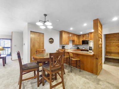 A kitchen and dining area with wooden cabinets, a round wooden dining table with four chairs, and modern lighting fixtures. The kitchen features a stove, oven, and microwave.