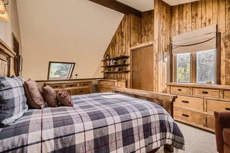 A rustic bedroom with wooden paneled walls, a bed with plaid bedding, and a skylight. The room features multiple dressers and a window with views of trees.