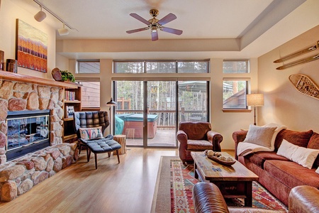 A cozy living room with a stone fireplace, sofa, armchairs, and a wooden coffee table. A ceiling fan is above, and large sliding doors open to a deck with a hot tub. Natural light fills the space.