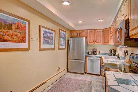 A small kitchen with wooden cabinets, stainless steel appliances, tile countertops, and framed art on the wall. The room is well-lit with ceiling lights.