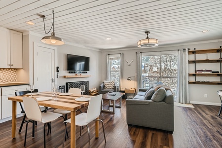 A modern, open-plan living and dining area with a wooden dining table, white chairs, grey couch, wall-mounted TV, and large windows letting in natural light.