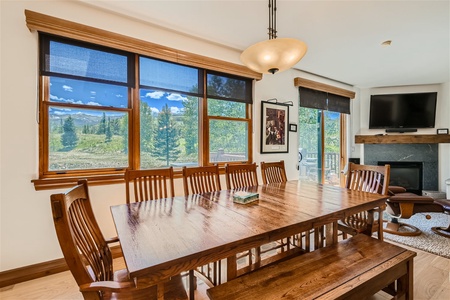 A dining room with a long wooden table and matching chairs. Large windows provide a scenic outdoor view. The room includes a ceiling light, a wall-mounted TV, and a fireplace in the corner.