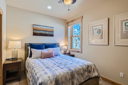 A cozy bedroom featuring a neatly made bed with blue and white bedding, nightstands with lamps on either side, artwork above the bed, and a window allowing natural light into the room.