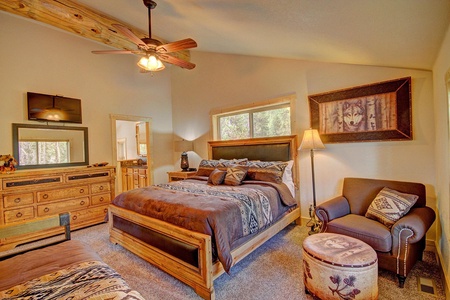 A rustic bedroom with wood furnishings, including a bed, dresser, and armchair. The room features a ceiling fan, a window, a wolf-themed wall picture, and a lamp beside the armchair.