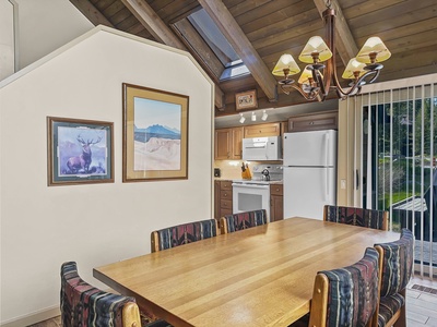 A dining area with a wooden table and eight upholstered chairs, an adjacent kitchen with appliances, and wall art featuring a deer and a landscape. A skylight and sliding glass door provide ample natural light.