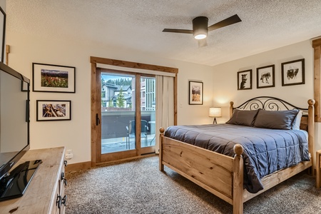 A well-lit bedroom with a wooden bed, gray bedding, wall-mounted TV, framed pictures on the walls, a ceiling fan, and sliding glass doors leading to a balcony.