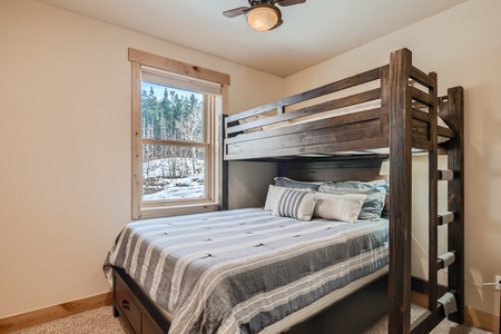 A bedroom with a dark wood bunk bed, twin on top and full-size on bottom, with striped bedding. A large window offers a view of a snowy landscape with trees. A ceiling fan and light fixture are visible.