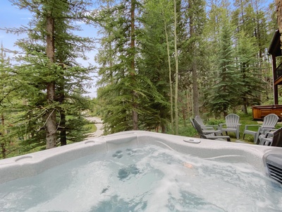 A bubbling hot tub overlooks a forested area with tall pine trees and a few Adirondack chairs placed nearby.