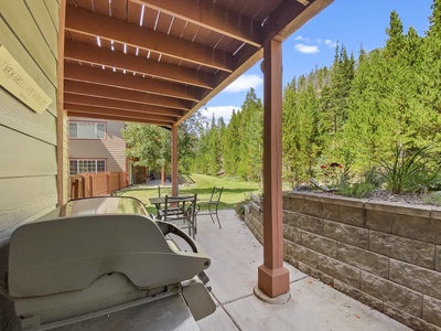 A covered patio with a grill, dining table, and chairs overlooks a lush, tree-filled backyard next to a residential building.