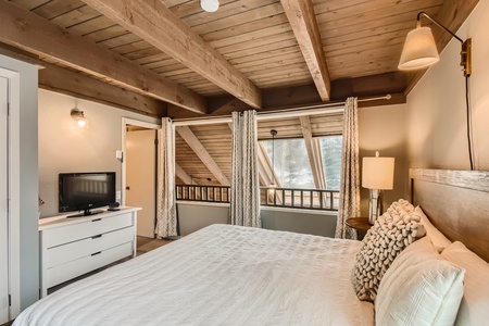 A cozy bedroom with wooden beams, a bed with white linens, a wall-mounted lamp, a small TV on a dresser, and a large window with curtains.