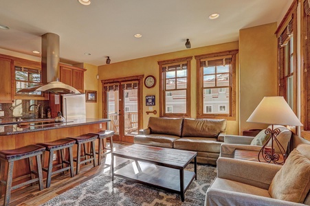 A cozy living room with a leather couch, armchair, coffee table, and a lamp. Adjacent is a kitchen with a breakfast bar, stools, and modern appliances. Sunlight enters through the windows and glass door.