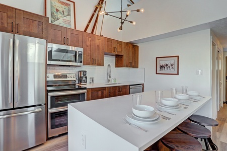 Modern kitchen with wooden cabinets, stainless steel appliances, and an island with bar stools. Table set with plates, bowls, and glasses. Bright lighting and framed artwork on the walls.