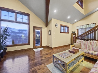 A living room with wood flooring, a striped sofa, a coffee table, and large windows. There are stairs to the right and a doorway leading outside. The ceiling has recessed lighting and visible beams.