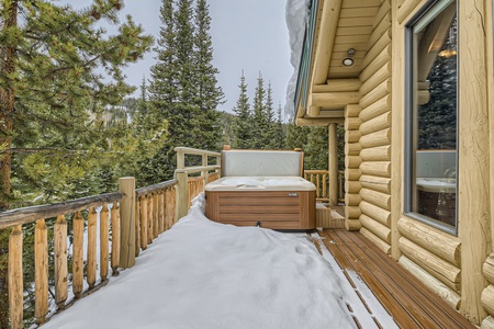 A wooden cabin with a snow-covered deck features a hot tub surrounded by pine trees.