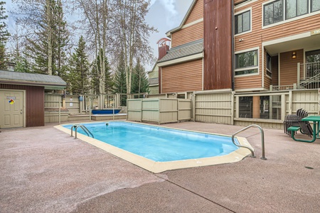 A small outdoor swimming pool with a metal ladder, surrounded by a fenced area and adjacent to a multi-story building with brown siding.