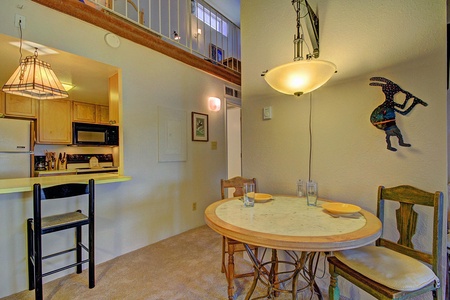 A small dining area with a round table and two chairs, adjacent to a kitchen with wooden cabinets and appliances. A decorative wall hanging is present above the table.