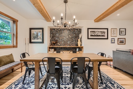 A dining room with a wooden table and six metal chairs. The room features wooden beams, a chandelier, framed art, a window seat, and a patterned rug.