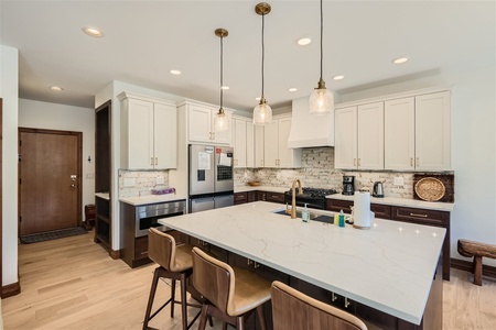 A modern kitchen with a large central island, marble countertops, light-colored cabinetry, three pendant lights, and stainless steel appliances. Four wooden barstools are placed around the island.