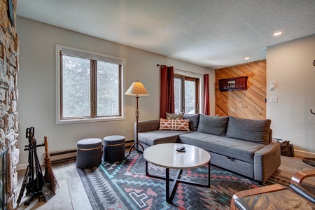 A cozy living room with a gray sectional sofa, round coffee table, patterned rug, floor lamp, and two ottomans, featuring a stone fireplace and large windows.