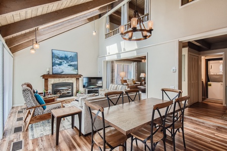 A living area featuring a wooden dining table with chairs, a seating area with armchairs and a couch, a fireplace, a wall-mounted TV, and an adjacent laundry area. Ceiling is wooden with exposed beams.