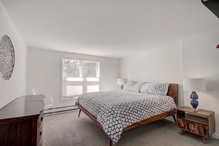 A minimalist bedroom with a double bed, two nightstands with lamps, a chest of drawers, a chair, and a window. White walls and carpet create a clean, bright atmosphere.