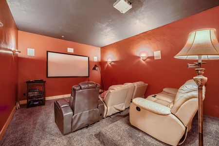 A home theater room with red walls, a large screen, a projector, and several recliner chairs arranged in rows. A floor lamp is visible to the right.