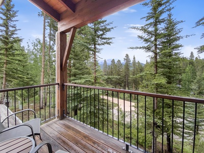 A wood balcony with metal railings overlooks a forested area with tall trees and mountains in the background under a clear sky. There are two chairs on the balcony.