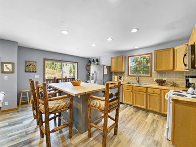 A modern kitchen with wooden cabinetry, a central island with rustic barstools, stainless steel appliances, and large windows providing natural light.