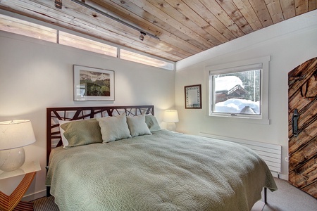 A cozy bedroom with a wooden ceiling, a large bed with green bedding, two table lamps, a wall mirror, and a window offering a snowy outside view.