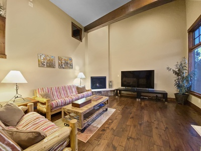 A living room with high ceilings, featuring a striped sofa, a coffee table, a TV, and a fireplace. The floor is wooden, and the room is lit by floor and table lamps. A plant is near the window.