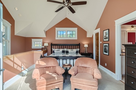 Spacious bedroom with slanted ceilings, featuring a bed, two beige armchairs, and a dresser. The walls are painted in a warm brown hue, and natural light illuminates the room through multiple windows.